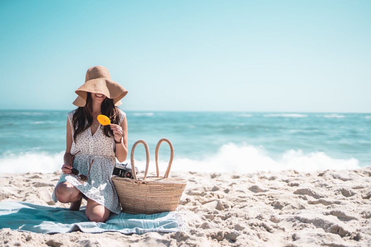 Woman in a sun hat enjoying a beach picnic with ice cream and camera on a sunny day.