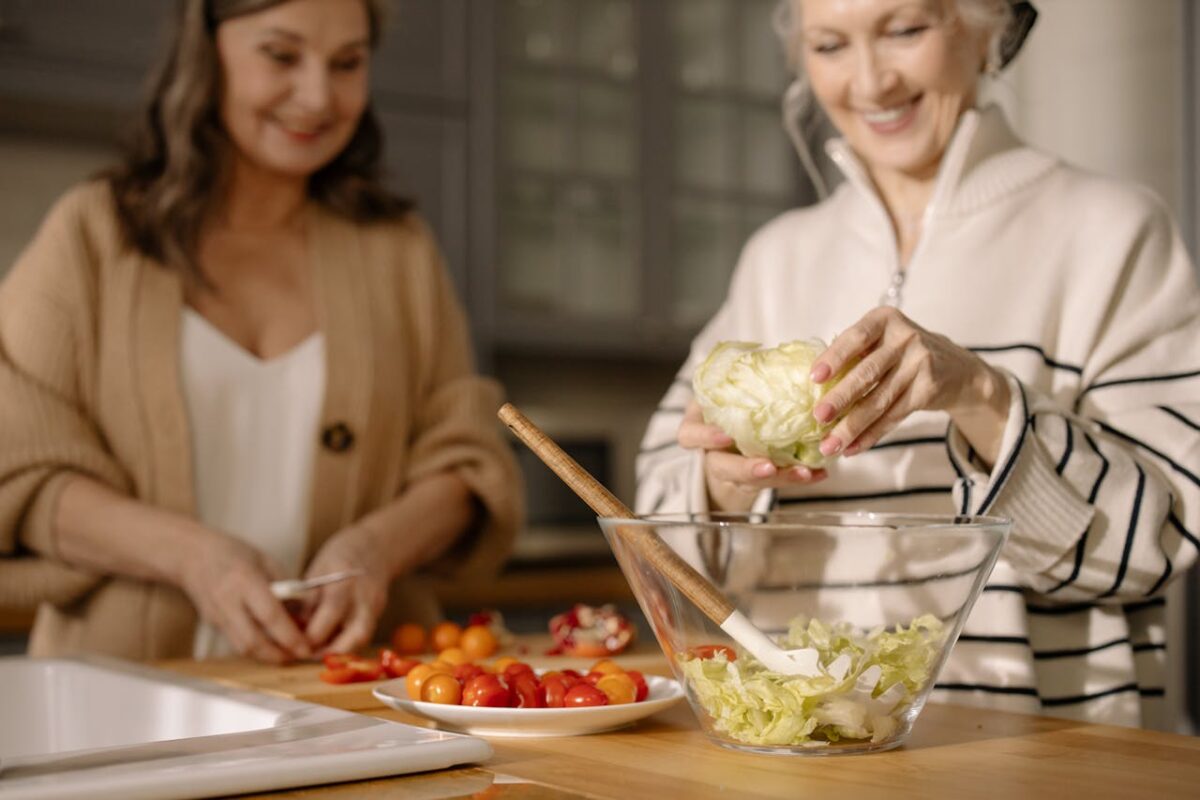 Senior women happily preparing a fresh salad with cherry tomatoes and lettuce in a cozy kitchen setting.
